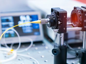 Testing device in an optical laboratory with a blurred display screen in the background, and light being shone through a yellow fibre optic cable.